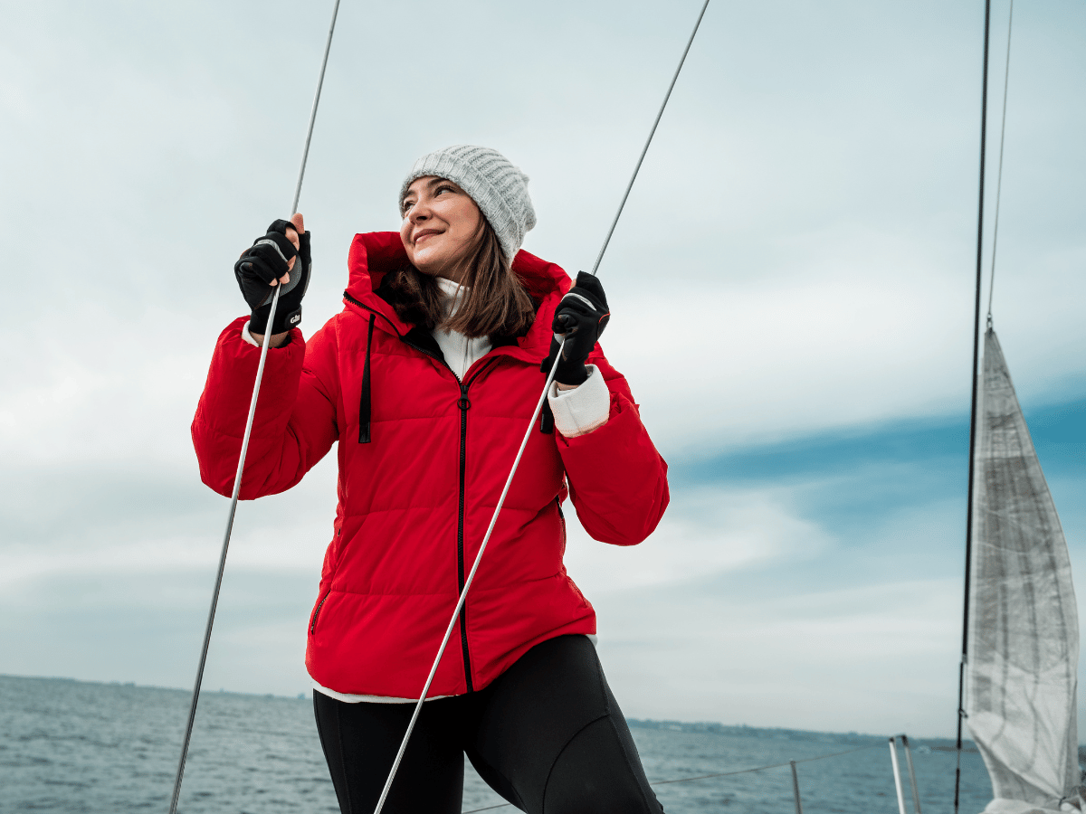 a woman in winter gear stands on deck with sails in the background